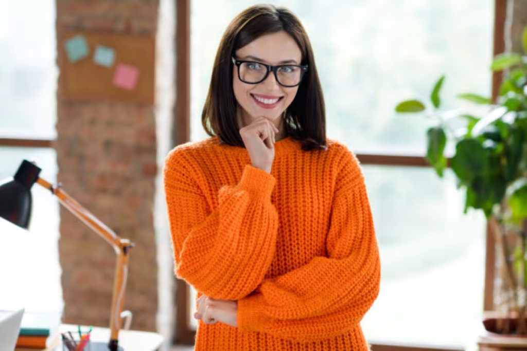 Smiling woman in an orange sweater and glasses with her hand on her chin